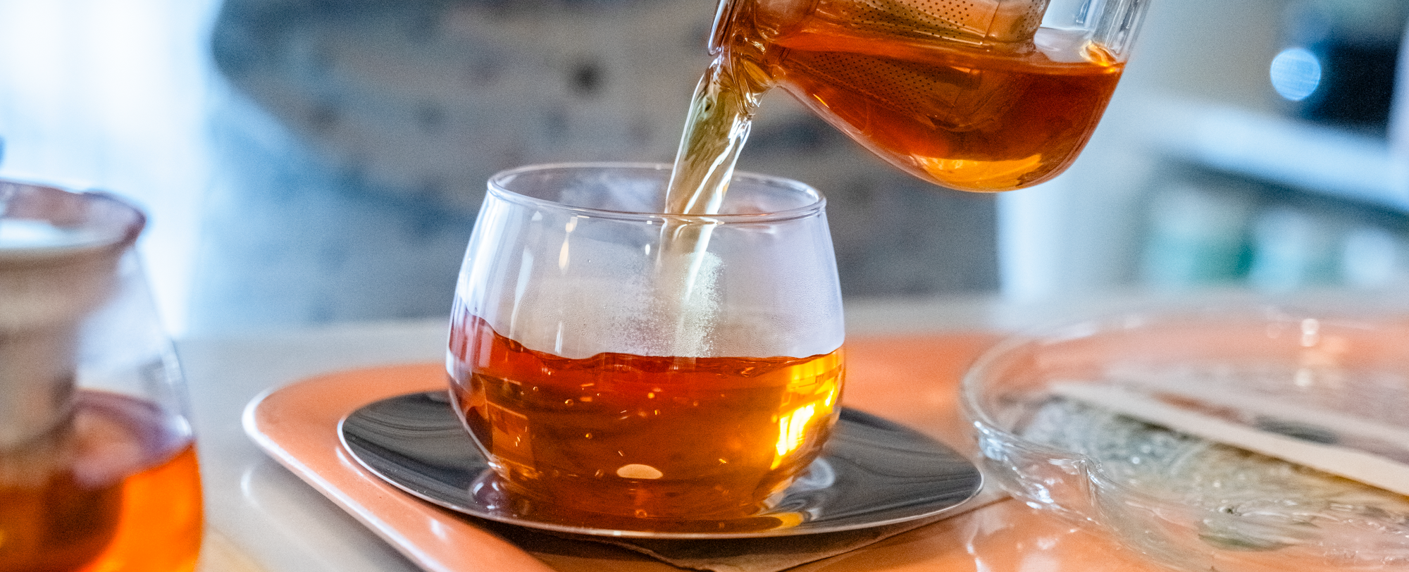 Person pouring tea from a glass teapot into a small glass cup on a table with blurred background